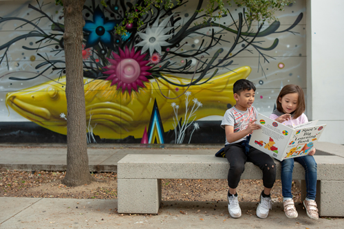 A first-grade boy reads a book to his kindergarten friend while sitting outside.