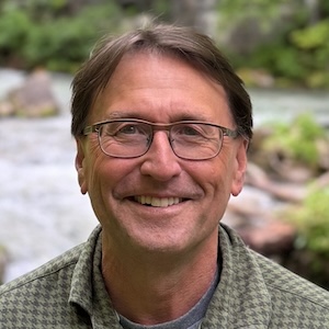 Roger Still wears glasses and a green checkered shirt, and smiles in front of a forest creek.