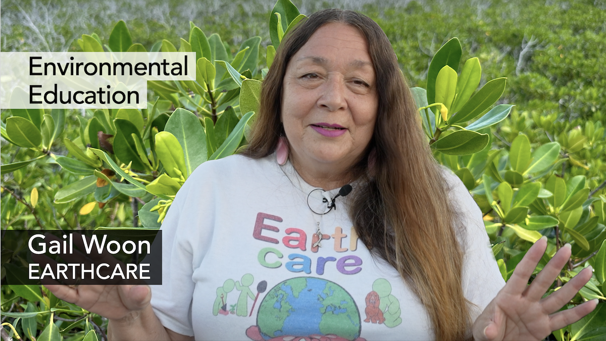 Gail Woon wearing a shirt that says "Earth Care" and standing in front of bright green leaves