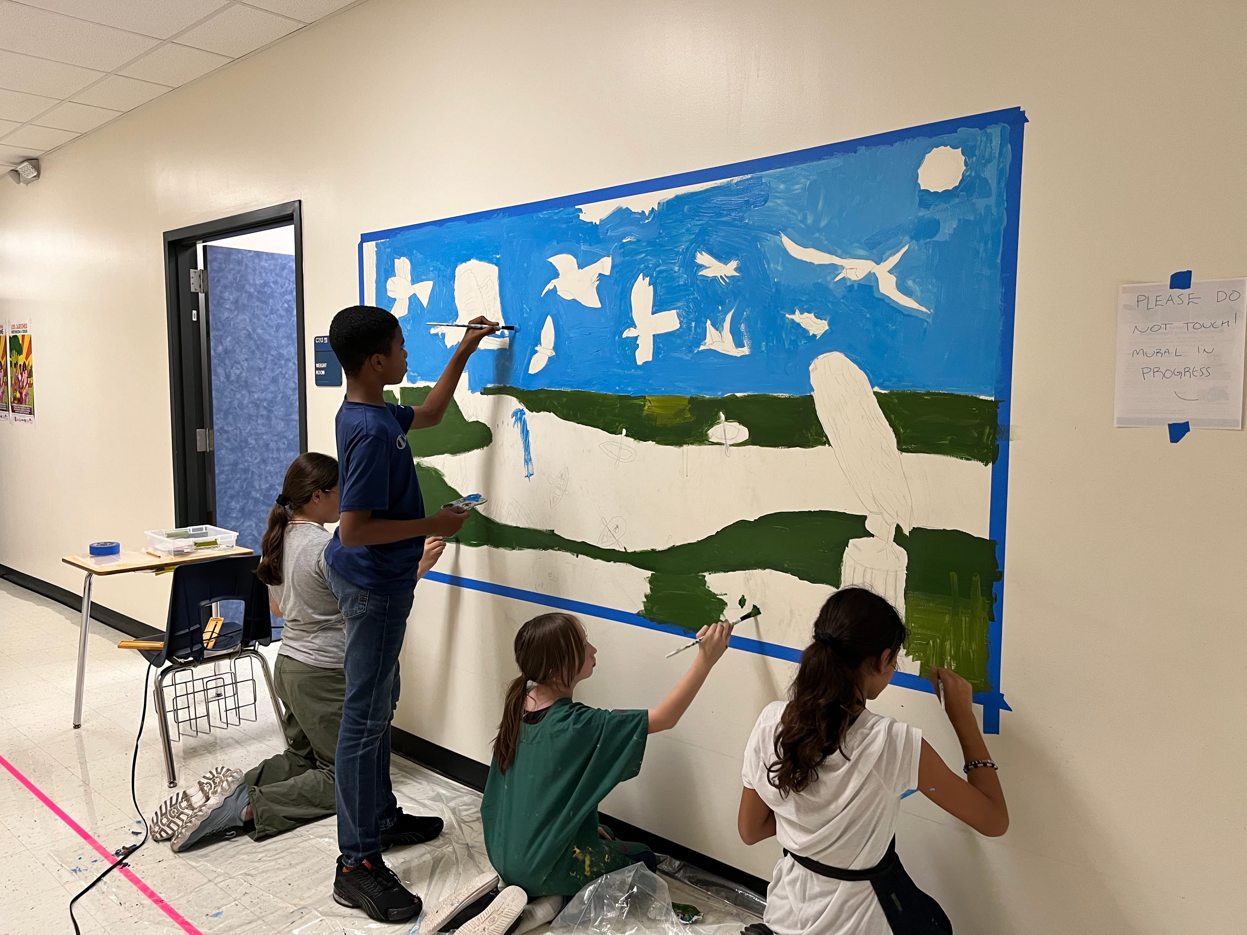 four elementary students paint a mural of flying birds on a school hallway wall using greens and blues.