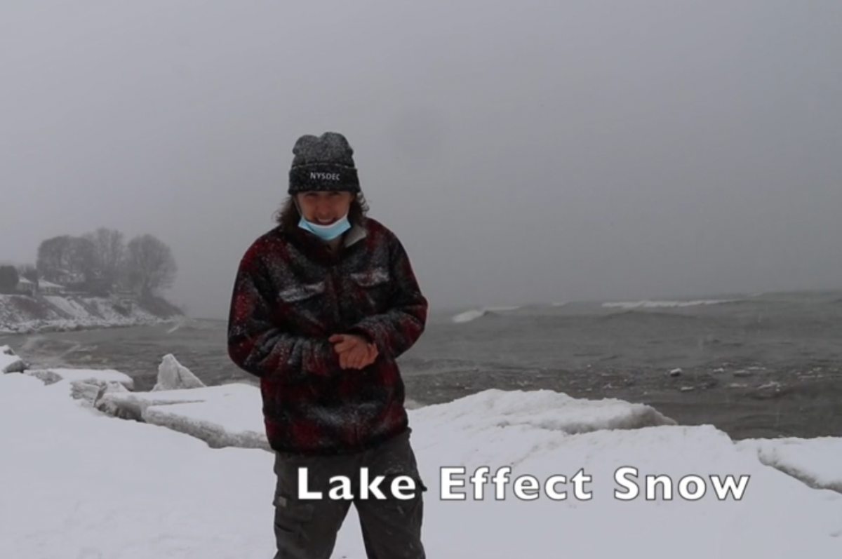 A man wearing beanie and thick plaid jacket standing outside on a snowy, windy day and near a lake.