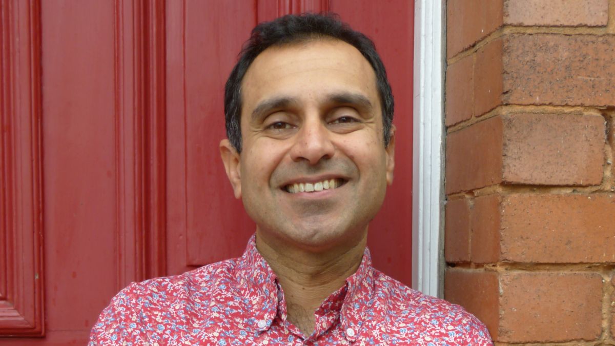 Portrait of Dr. Vik Mohan smiling and in a red, long-sleeve button up shirt, leaning against a brick exterior and in front of a red door
