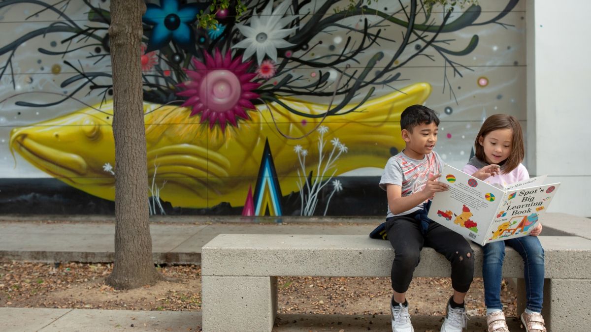 A first-grade boy reads a book to his kindergarten friend while sitting outside.