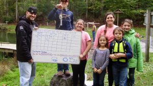 A photo of a family holding a whiteboard and thermometer in a forest clearing.