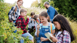 Group of students outside using a magnifying glass and a notebook to record what they see in a shrub