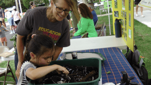 CEE-Change Fellow Melanie Schikore engages children at the farmer's market. Melanie is smiling while one child is focused and picking up worms from soil in the green worm bin.