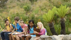 five elementary students writing on clipboards and sitting on rock wall in front of green shrubbery