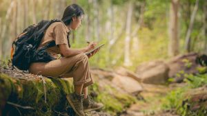 Person wearing backpack, sitting on mossy stones in forest to take some notes.