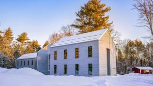 A light-gray building with an A-frame roof stands prominently in morning sunlight. A layer of snow sits on the roof and deep snow is on the ground. Behind the building are leafless trees and conifers, lit brightly by yellow morning sunlight. Above the trees is blue sky.
