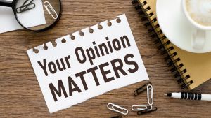 "Your opinion matters" written on notebook paper, sitting on wooden table next to notebook, coffee, paper clips