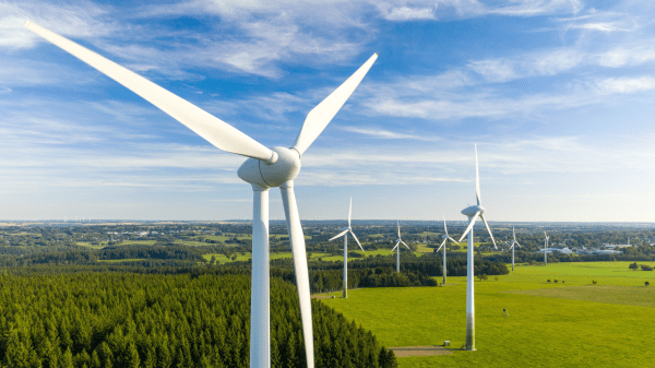 Wind turbines across a rural field; behind them is a forested area and a lightly cloudy blue sky