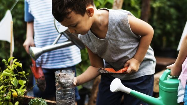 Three children around a boarded garden bed, two holding watering cans and one holding a plastic bottle with dirt