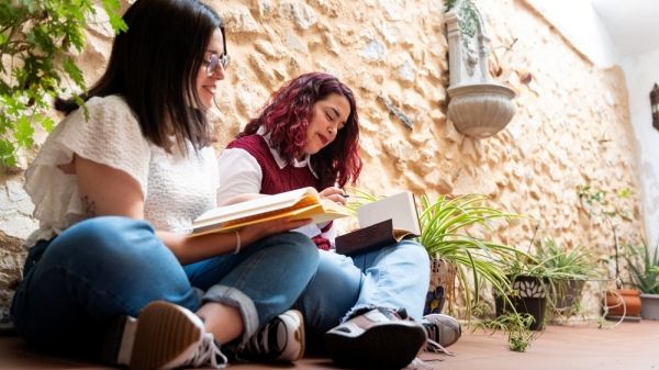Two women site side by side reading books outside against a wall.