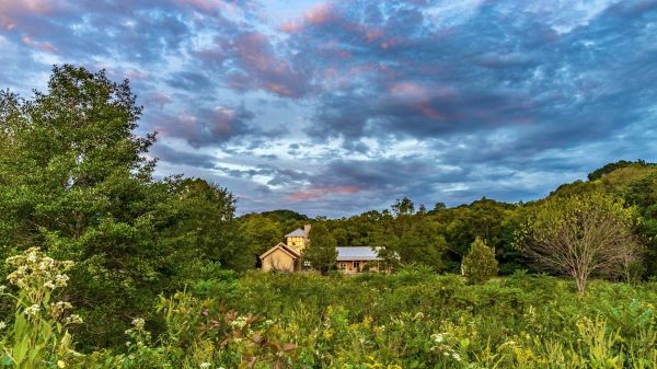A building stands in the distance, surrounded by a prairie in the foreground, forested hills behind, and a stunning blue sky above.