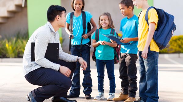 Teacher crouching next to four elementary aged students who are smiling and wearing backpacks.