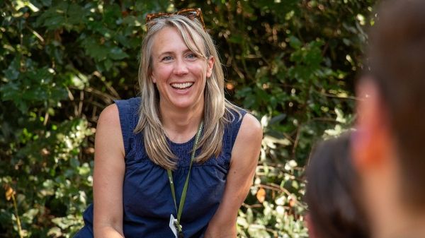 A long-haired person smiles widely at two people in the foreground. Behind them, foliage speckled with sunlight.