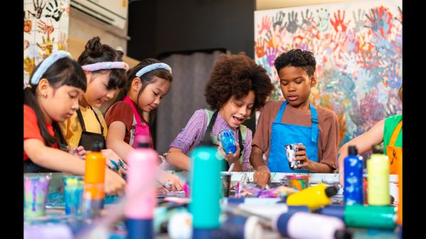 Give an Hour for Earth: World Water Day Youth Art Gallery Blurred paint bottles sit in front of five children all painting around a table; the back wall features paint handprints"