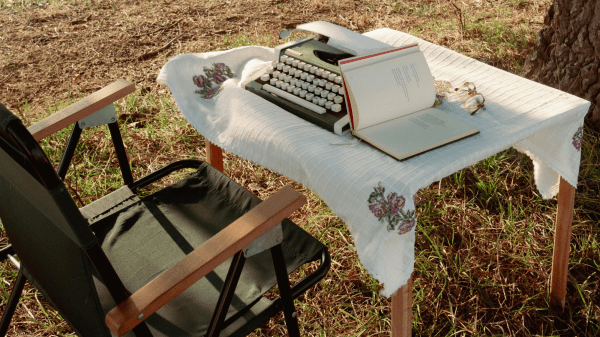 Author Talks A green folding chair sits to the left of a table with a white cloth on it; on top of the table is a green typewriter and open book; behind this the ground is covered with yellow grass