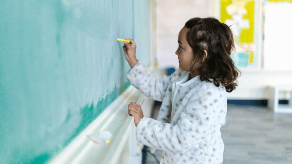 Child Chalkboard A child writes on a green chalkboard with a piece of chalk held in their right hand; behind them is a classroom wall