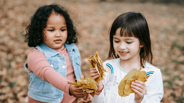 Children and Nature Two children hold clumps of brownish orange leaves, behind them is a blurred background where leaves can be seen on the ground