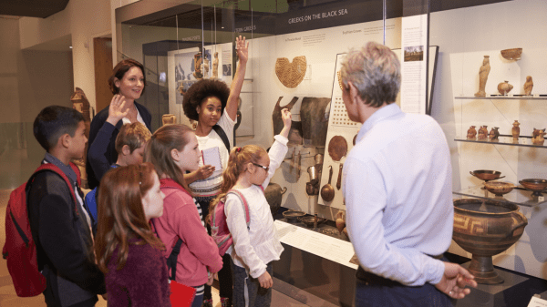 Children Museum A group of five children stand in front of an adult, three raising their hands; to the right is an adult facing them; behind all of them is a display of fossils