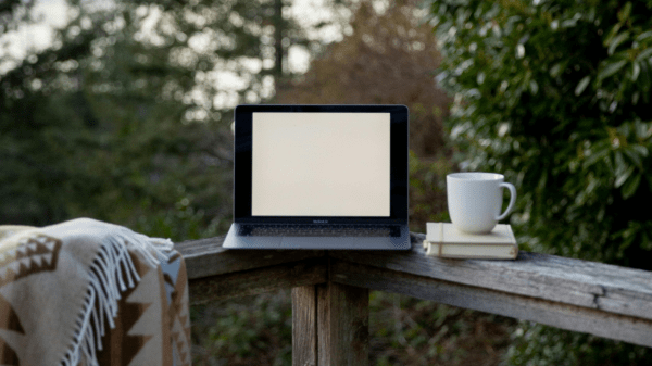 Computer and Nature An open laptop sits on a patio fence between a blanket and a coffee mug; behind it is a forest of trees