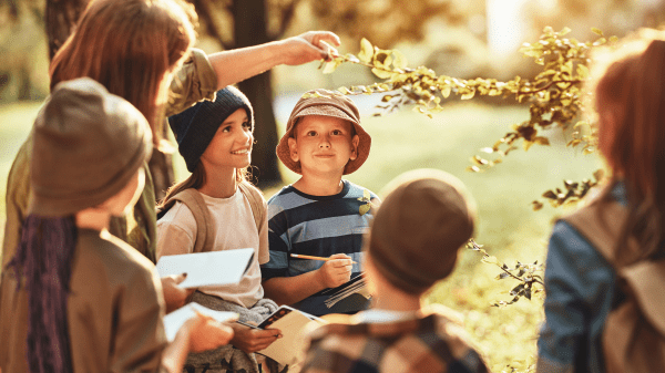 Nature School A group of children stand around an adult pointing to a leafy branch of a tree; behind them is a blurred grassy and tree-filled background