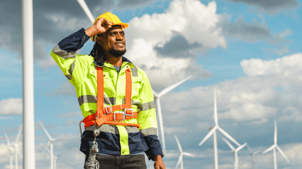 Green Careers Hub A person in a yellow visibility shirt to the left has a hand up to their hardhat; behind them is a cloudy sky with a series of wind turbines in front of it