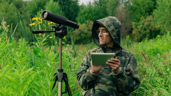 Green Jobs for Nature A telescope stands to the left of a person in a camo hood holding a clipboard; behind them is a field of grass with trees further in the distance