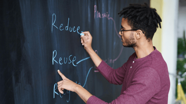 Sustainability Teaching A person writes "Reduce, Reuse, Recycle" in blue chalk on a chalkboard to the left of them; their left hand points to the word Reuse