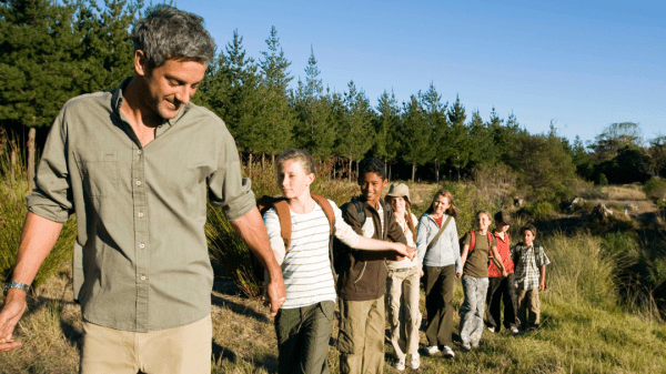 A teacher leads a group of children single file along a trail near a forest. The children follow closely behind. The trail is surrounded by lush greenery, and the forest can be seen in the background