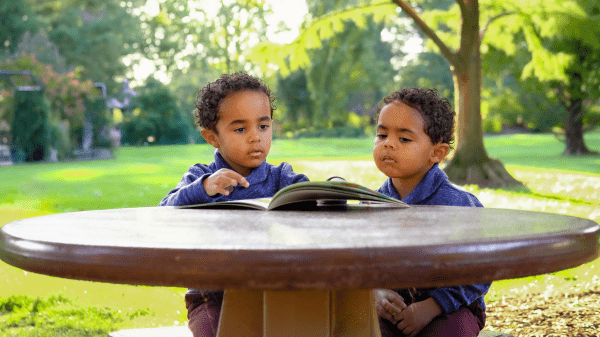Nature-Based Early Childhood Program Assessment & Guidebook Two children sit at a wooden table with a book in front of them and a grassy field and trees behind them