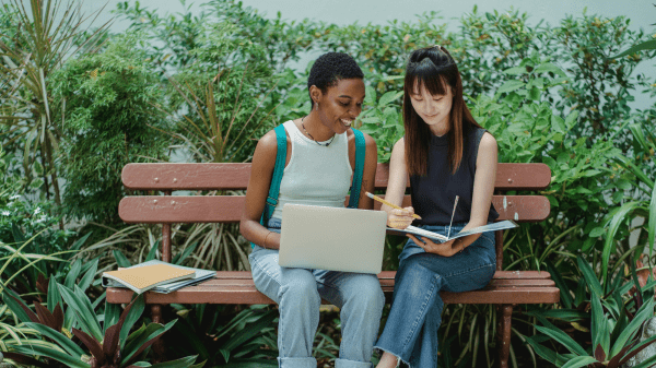 Nature-Based Teacher Certification (Online) Two people sit on a park bench with notebooks to the left of them; the person on the left has a laptop in their lap, the person on the right is holding a notebook and pencil; behind them is an assortment of green foliage
