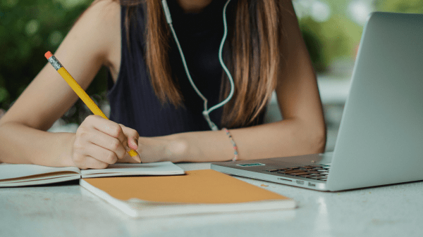 Certificate Course A person, head cut off by the image ending at the top, sits at a table outside with a laptop on it; they're wearing earbuds and holding a pencil over some paper and a notebook