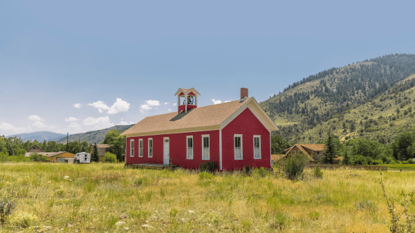 Nine Action Steps That Build Rural School Sustainability A red schoolhouse sits in the center of a green grassy field, behind it is a green tree-dotted hill and a slightly cloudy blue sky