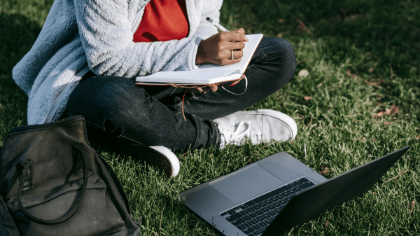 Online Outdoor Courses A person holding a notebook and pen sits to the left with their head cut off by the image ending; below them is a black bag and a laptop, both in the grass