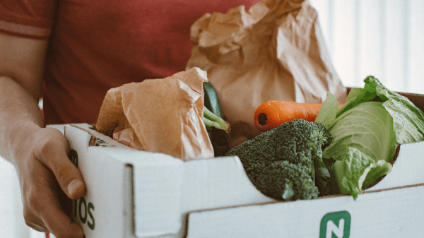 A person in a red shirt holds a cardboard box with vegetables
