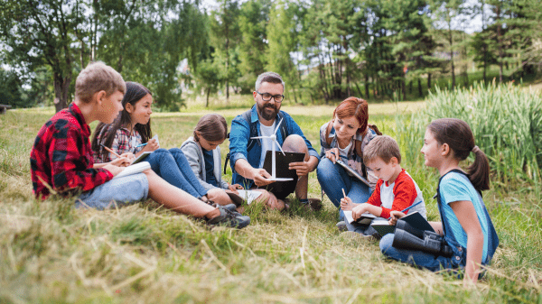 Reimagining the Role of Teachers in Nature-Based Learning: Helping Children Be Curious, Confident, and Caring Two adults sit in grass, one holding a miniature wind turbine, surrounded by children and all in a grassy field with trees further in the background