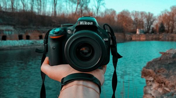 Photo Contest A hand holds out a camera facing the viewer, beyond it is a river