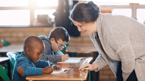 Teaching Sustainability in Rural US Classrooms Two students sit at their desks to the left of the image, to the right of them is an adult instructor standing and leaning over the desks; the background features two windows