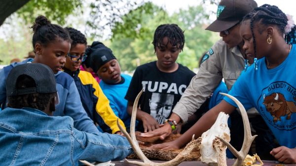 Students gather around a table outside surrounded by trees with outstretched hands to touch a variety of pelts and bones as part of an environmental education program led by a park ranger at a local park