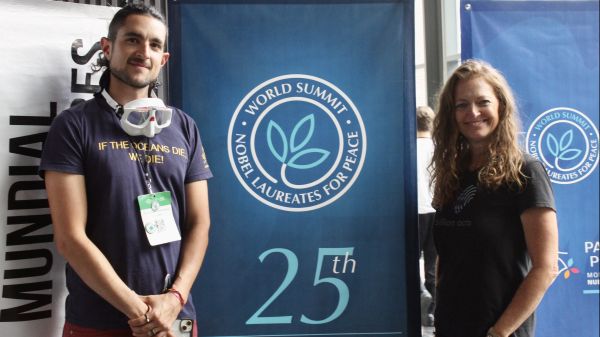 World Summit For Nobel Laureates For Peace Billion Acts of Peace Fellowship Mentor at Monterrey, Mexico 2024; two people stand on either side of a "25th Founding Anniversary, World Summit Nobel Laureates for Peace" banner