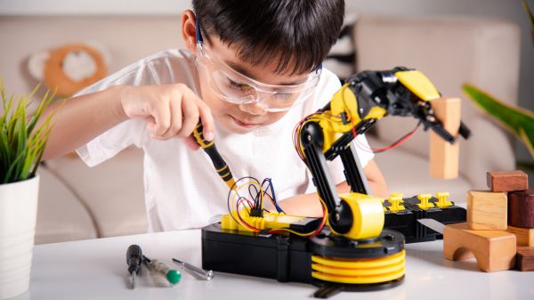 STEM Kids Work A child in a white shirt wearing safety glasses holds a screwdriver in their right hand; in front of them on a table is a yellow robotic arm