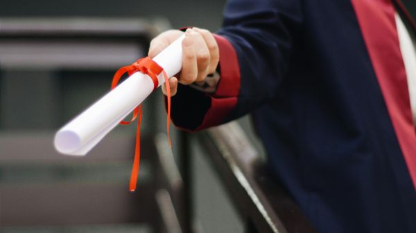Graduate Program A hand holds out a rolled piece of paper tied with an orange ribbon; the person is wearing a graduation robe