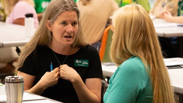 A woman in a black shirt faces another long-haired person, speaking and gesturing with her hands to herself.