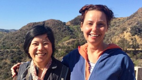 Climate Stories Ambassadors Two people stand next to each other smiling, behind them is a hilly landscape with a clear blue sky