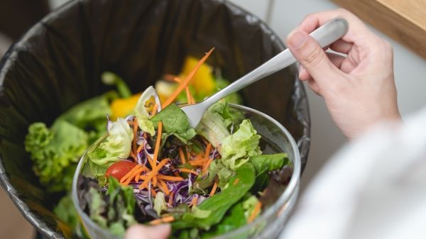 A person holds a salad bowl above a trashcan with other food waste in it.