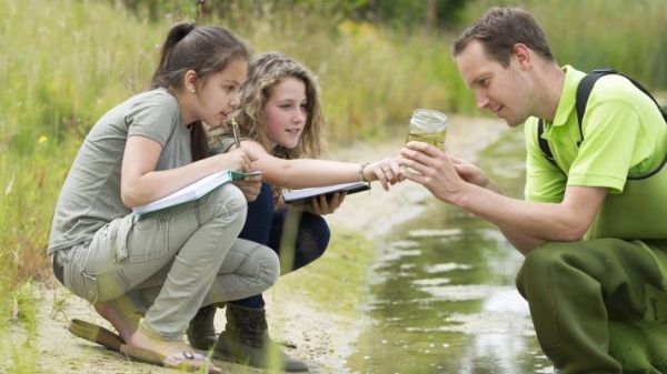 Group of three people near water. Two young women hold open notebooks while looking at a clear cup with water held by a young man.