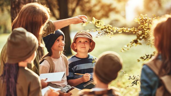 Group of curious school children with notebooks listening to their young female teacher while learning about nature together, looking at green leaf during ecology lesson in autumn forest on sunny day