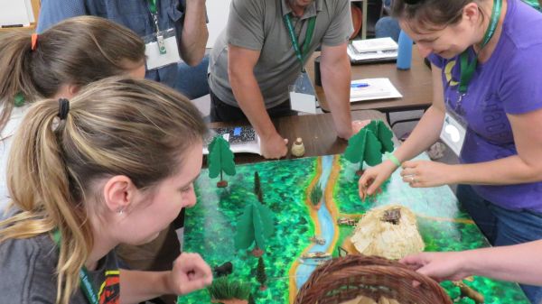 A group of five adults standing around a forested creek paper model.